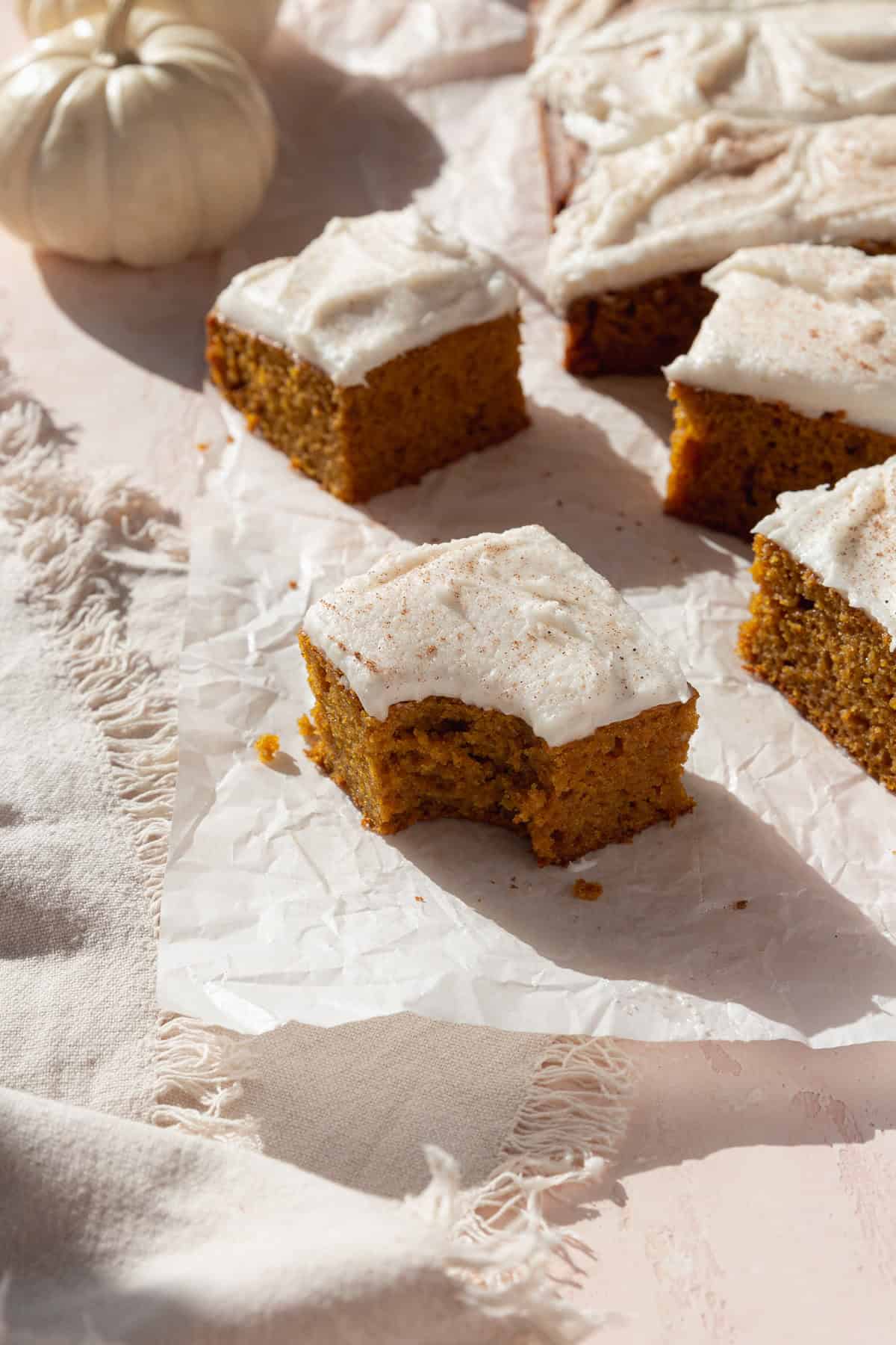 A bitten square of pumpkin bar with cream cheese frosting on a piece of parchment paper with more squares scattered behind.