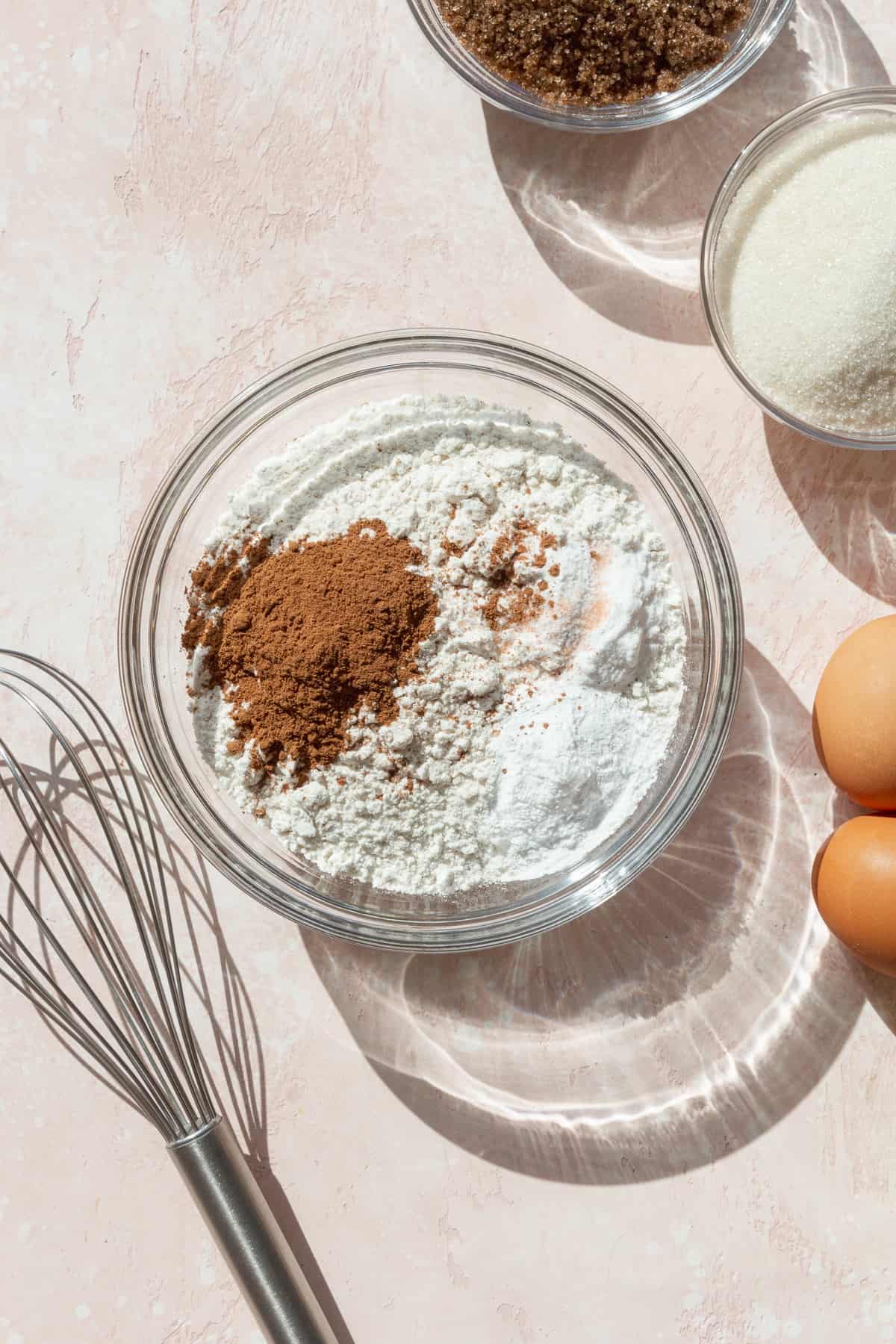 Flour, salt, baking soda, baking powder, and pumpkin pie spice in a glass bowl next to a wire whisk.