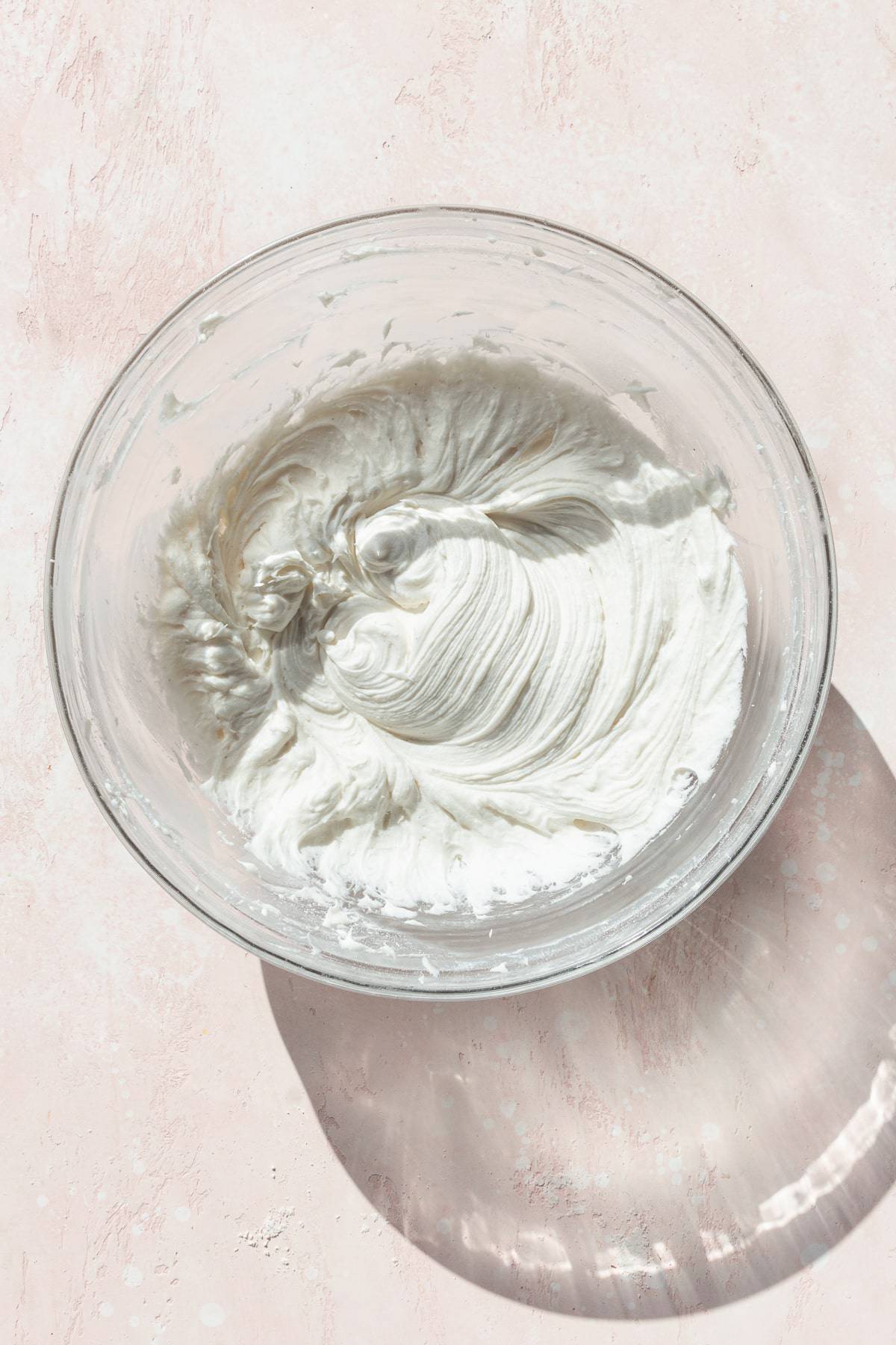 Cream cheese frosting in a glass bowl on a pink surface.