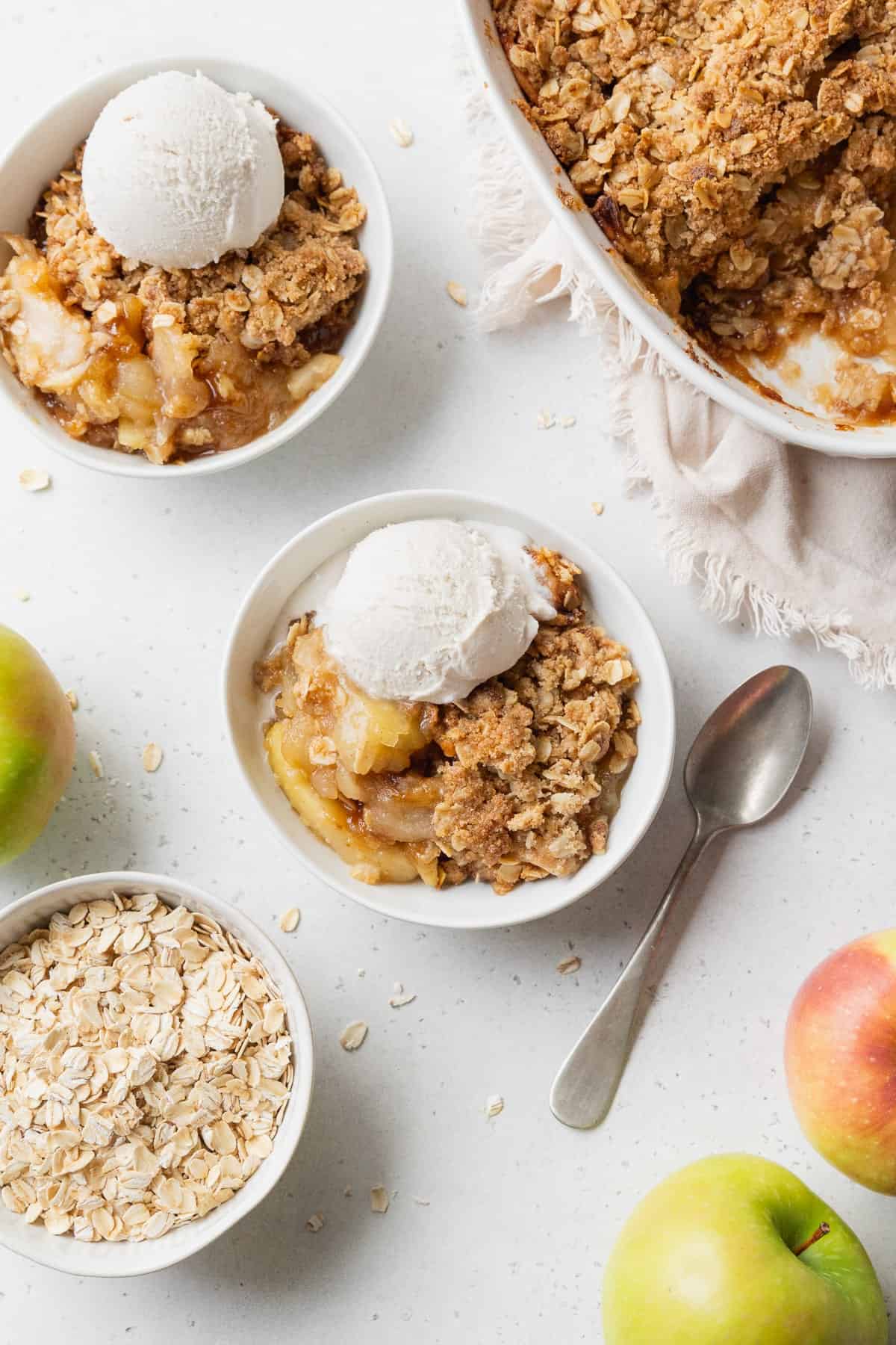 Two bowls of gluten free apple crisp with vanilla ice cream, and a baking dish of more apple crisp placed nearby.