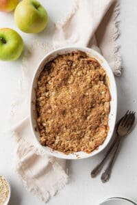 Baked apple crisp on a beige linen with a spoon, fork, and some green apples placed next to it.