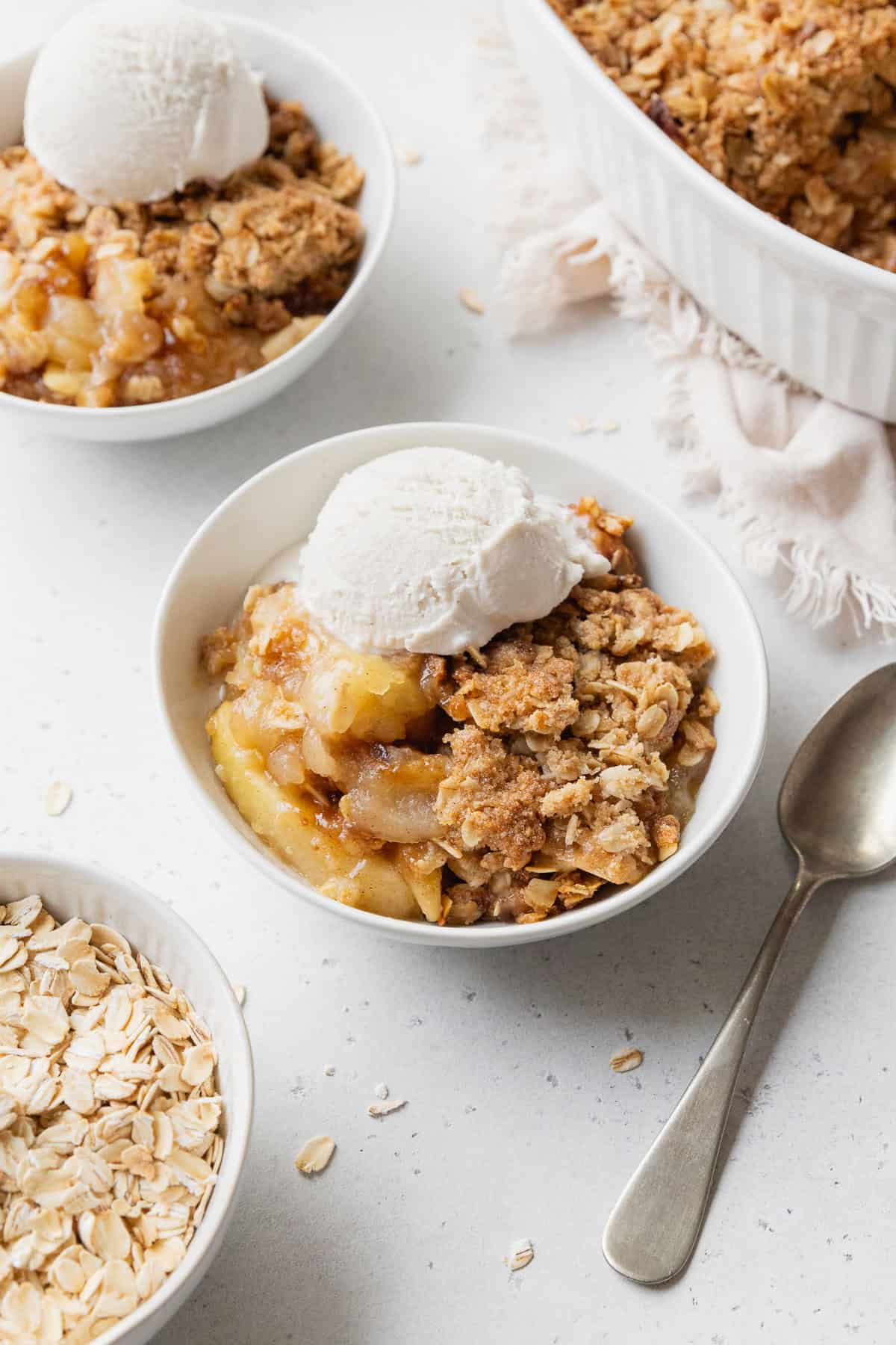 Gluten-free apple crisp with vanilla ice cream in a small white bowl next to a spoon with more bowls placed nearby.