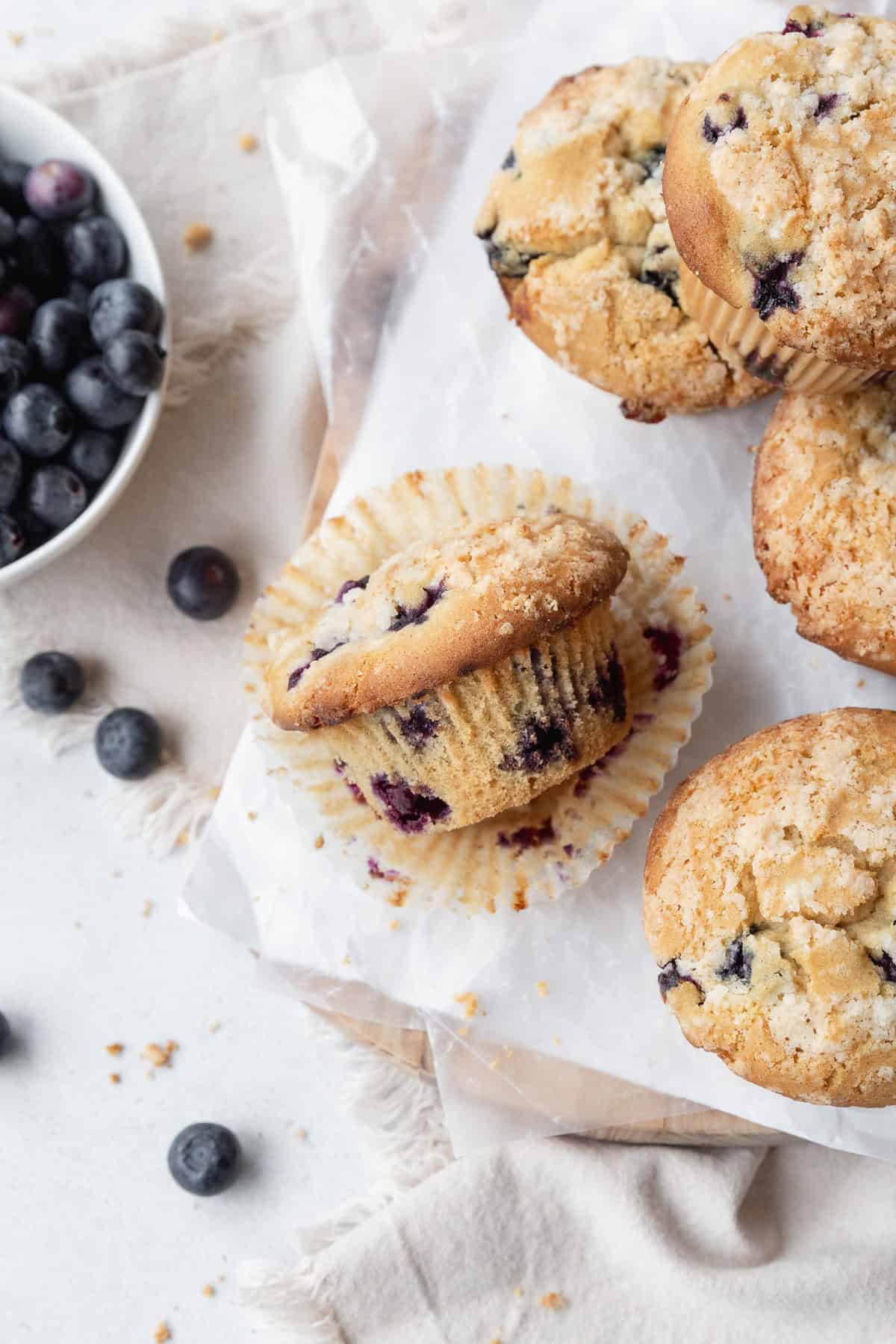 A blueberry muffin laying on its side on a paper liner, with more muffins on the right and a bowl of blueberries on the left.