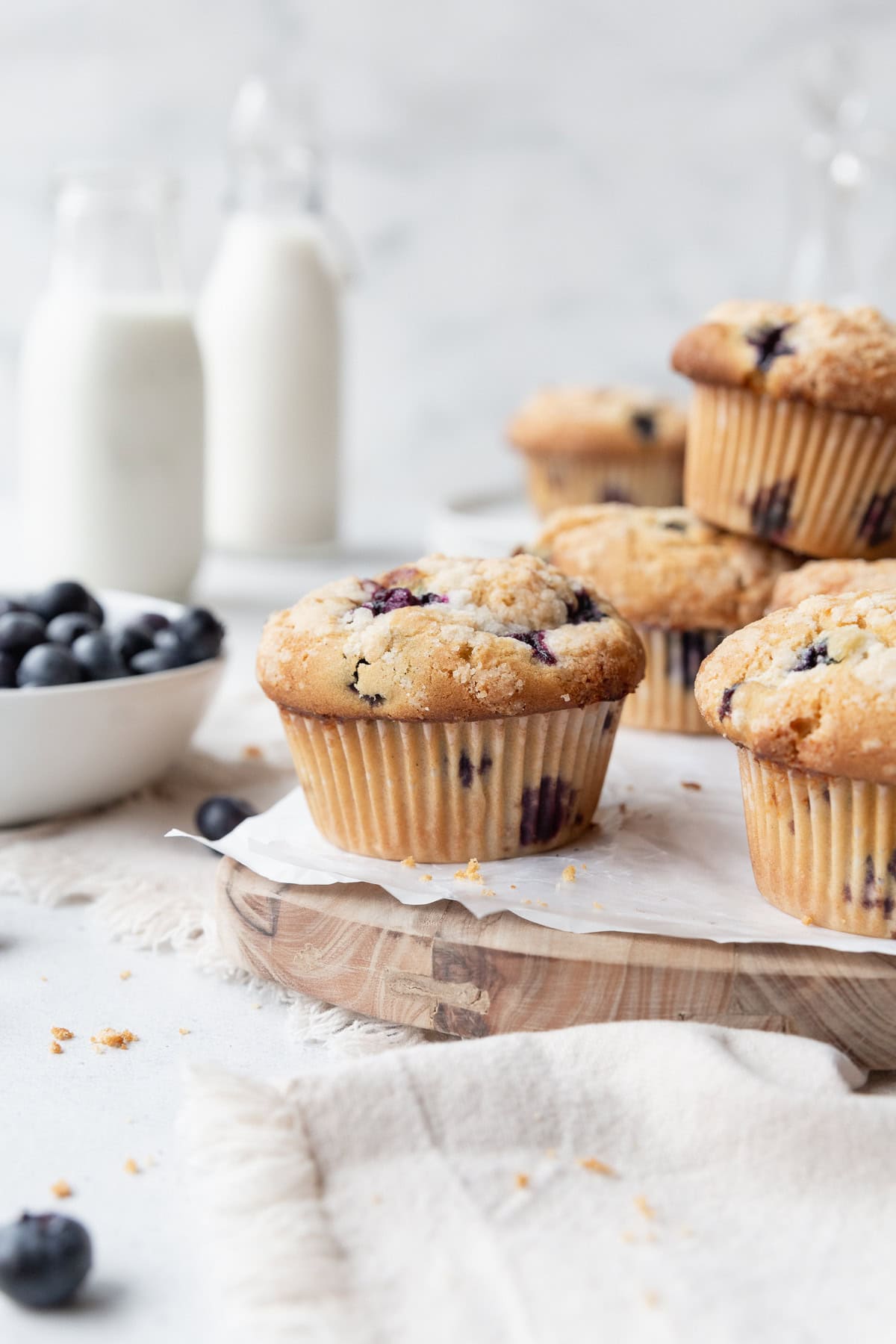 Gluten-free blueberry muffins on a wooden board lined with parchment paper, with a bowl of blueberries on the side and milk jars in the background.