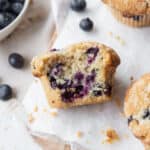 A bitten gluten free blueberry muffin laying on its side on parchment paper.