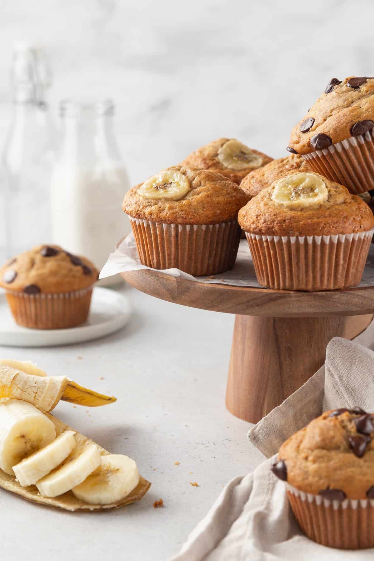 Gluten-free banana muffins on a wooden cake stand, with some sliced bananas on the surface underneath, and glass milk bottles in the background.