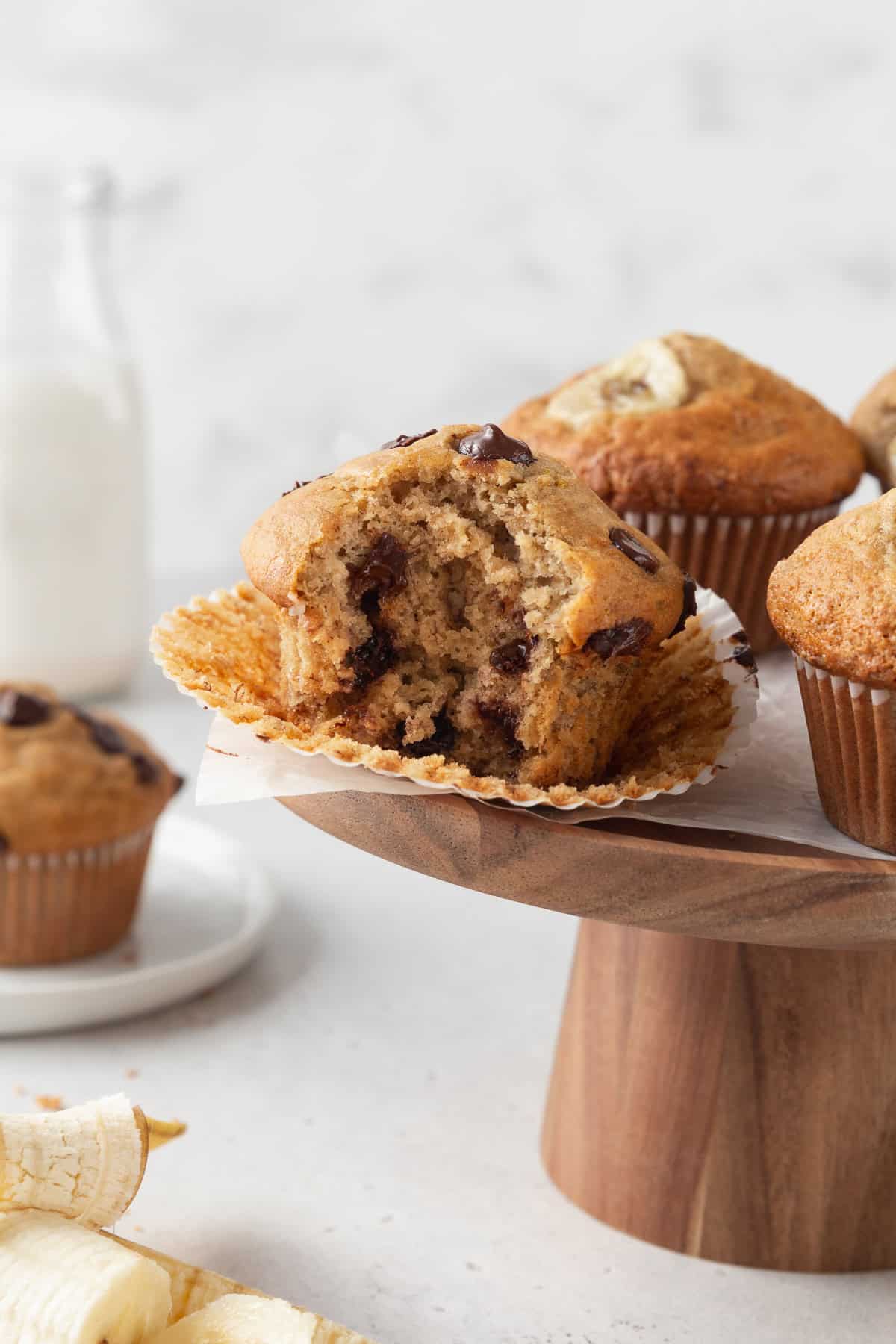 A bitten gluten free banana chocolate chip muffin on a wooden cake stand.