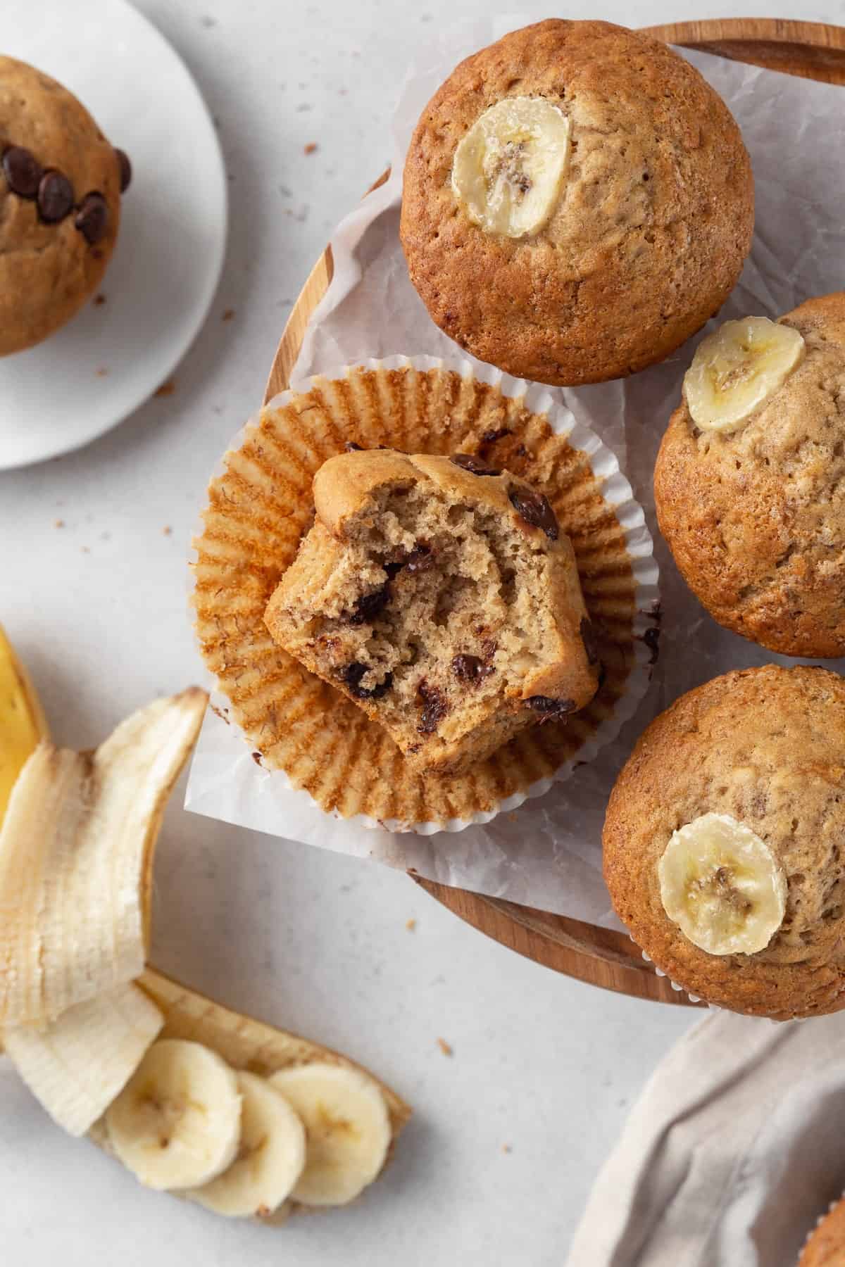 A bitten chocolate chip banana muffin on a paper liner surround by three more muffins.