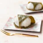 Sakura mochi wrapped in a cherry blossom leaf on a square pink plate with a gold fork laying in front.