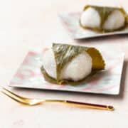 Sakura mochi wrapped in a cherry blossom leaf on a square pink plate with a gold fork laying in front.