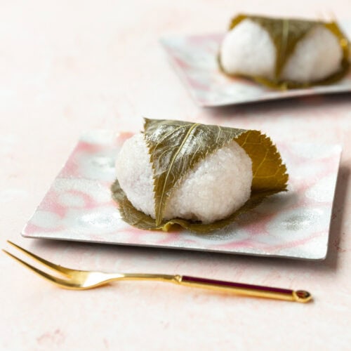 Sakura mochi wrapped in a cherry blossom leaf on a square pink plate with a gold fork laying in front.