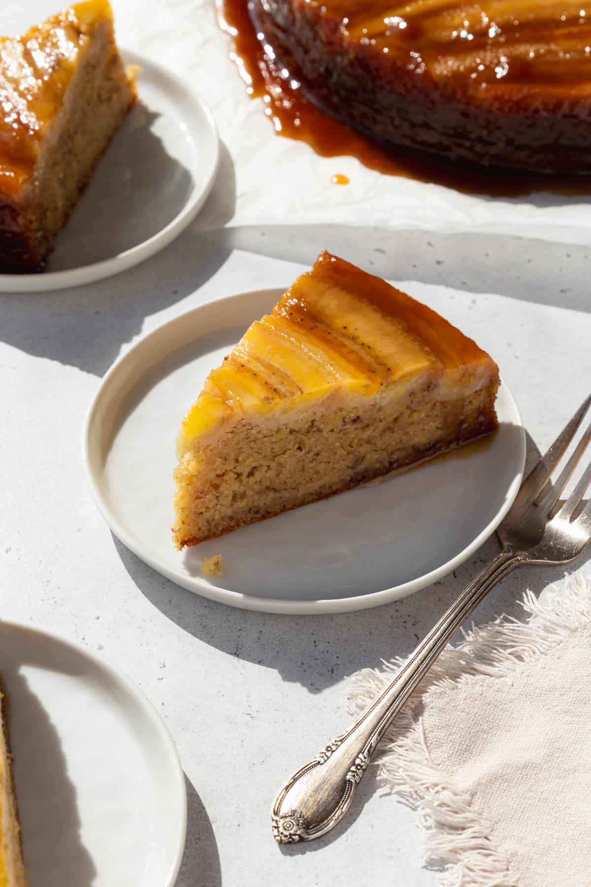 A slice of upside down banana cake on a white dessert dish with a silver fork next to it.