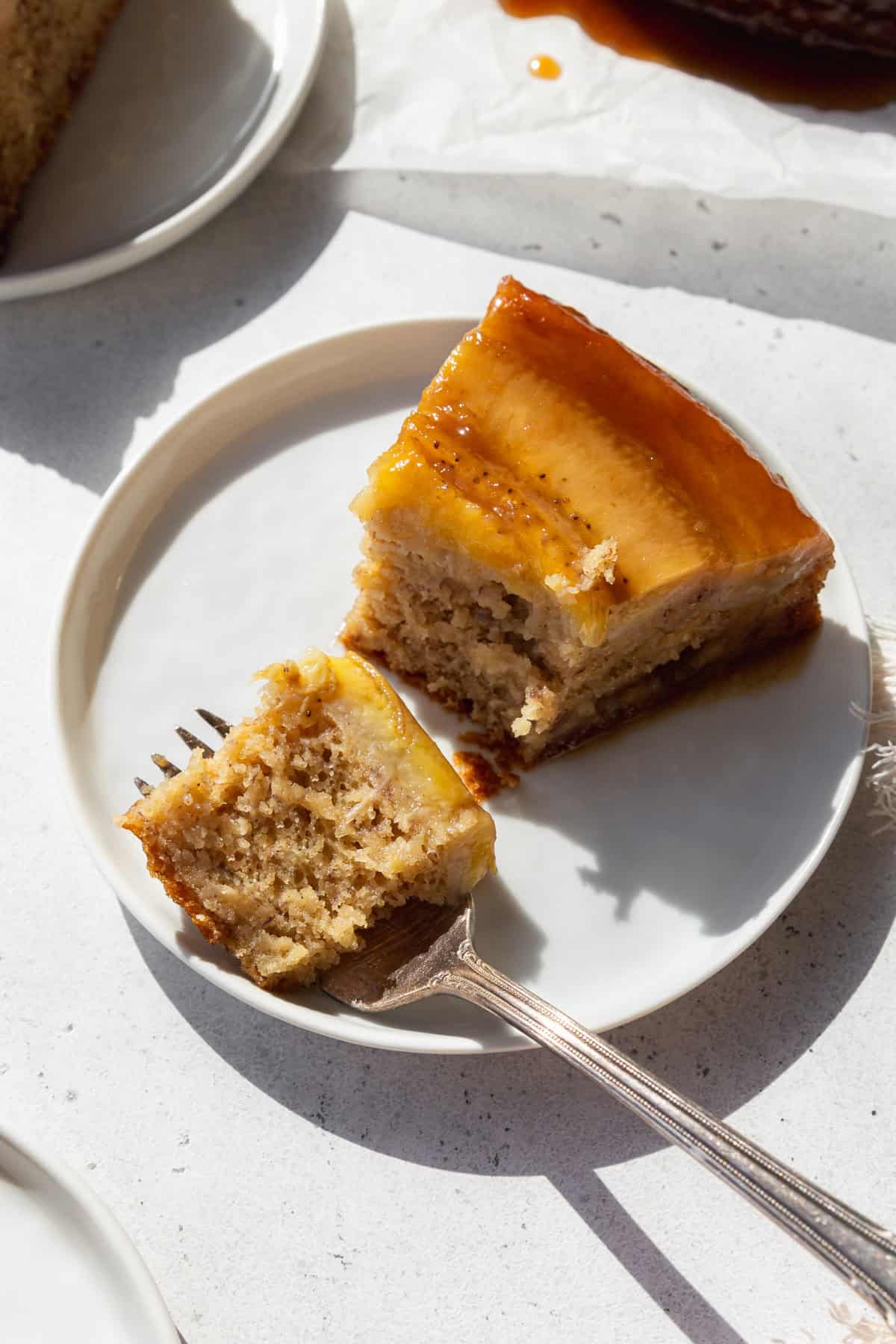 A slice of banana upside down cake on a white plate with a fork holding a bite of cake in front.