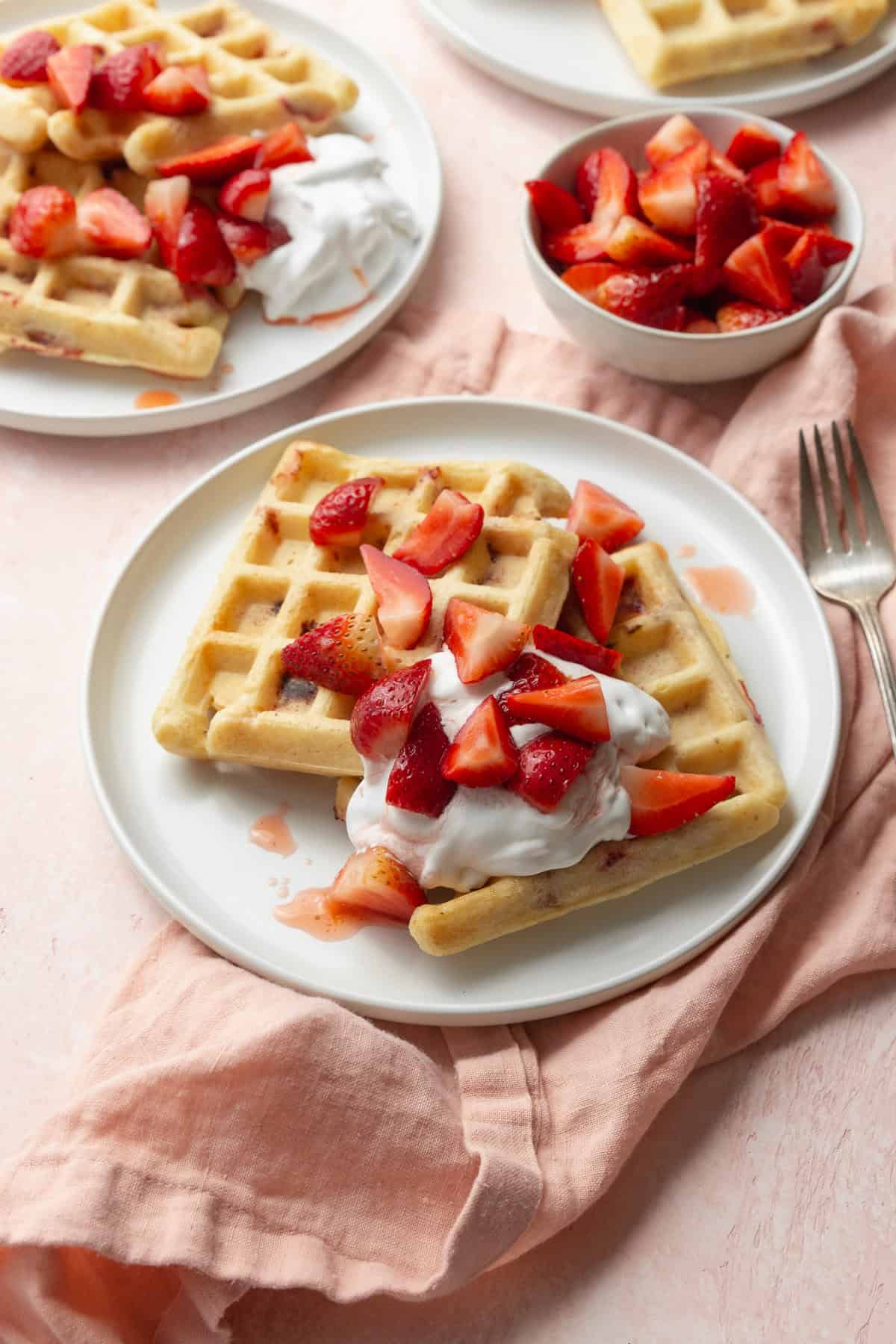 Strawberry waffles on a white plate with a dollop of whipped cream and sliced strawberries, with another plate of waffles and a small bowl of strawberries in the background.