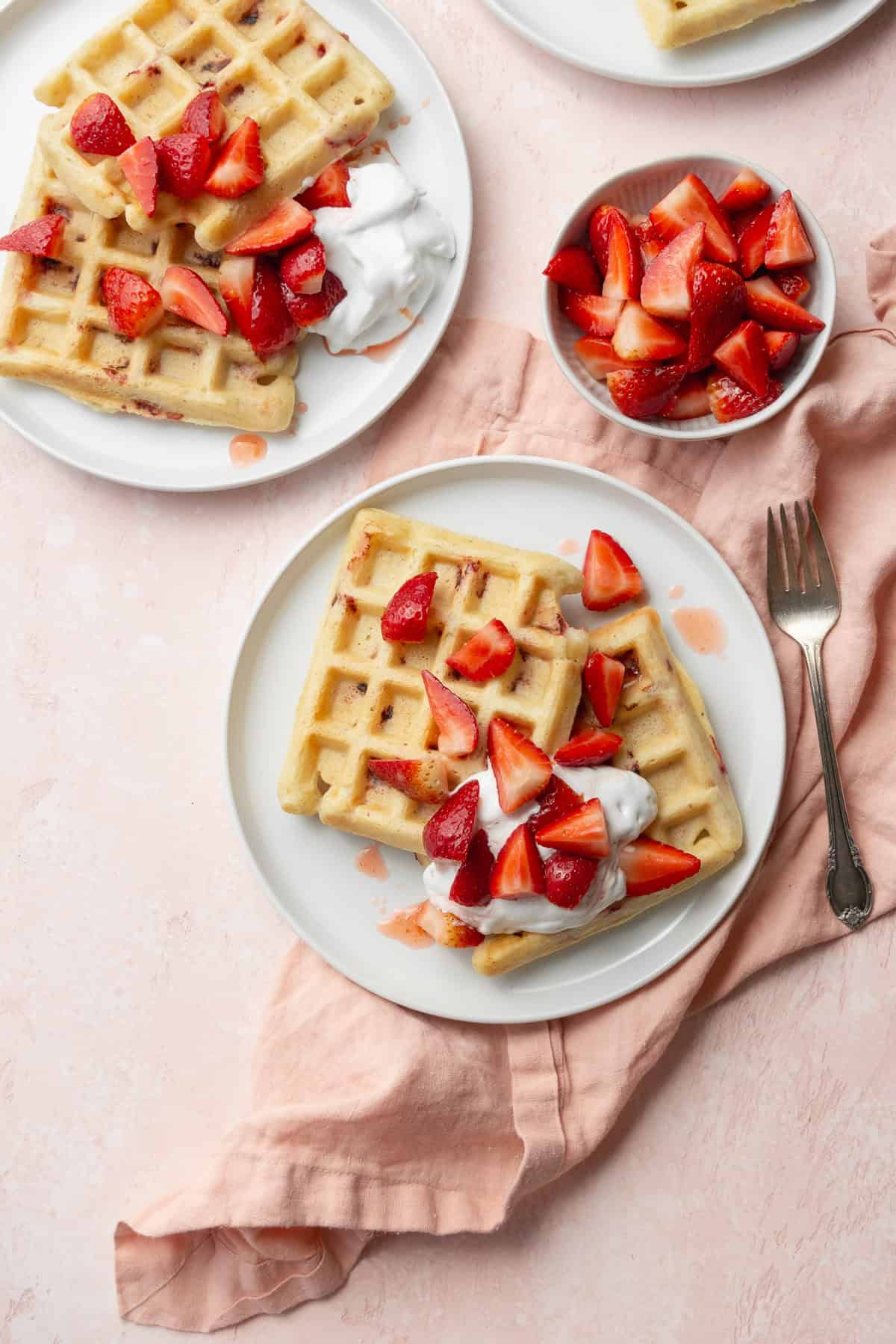 Two plates of waffles with strawberry topping next to a small bowl of strawberries on a pink surface with a pink napkin.