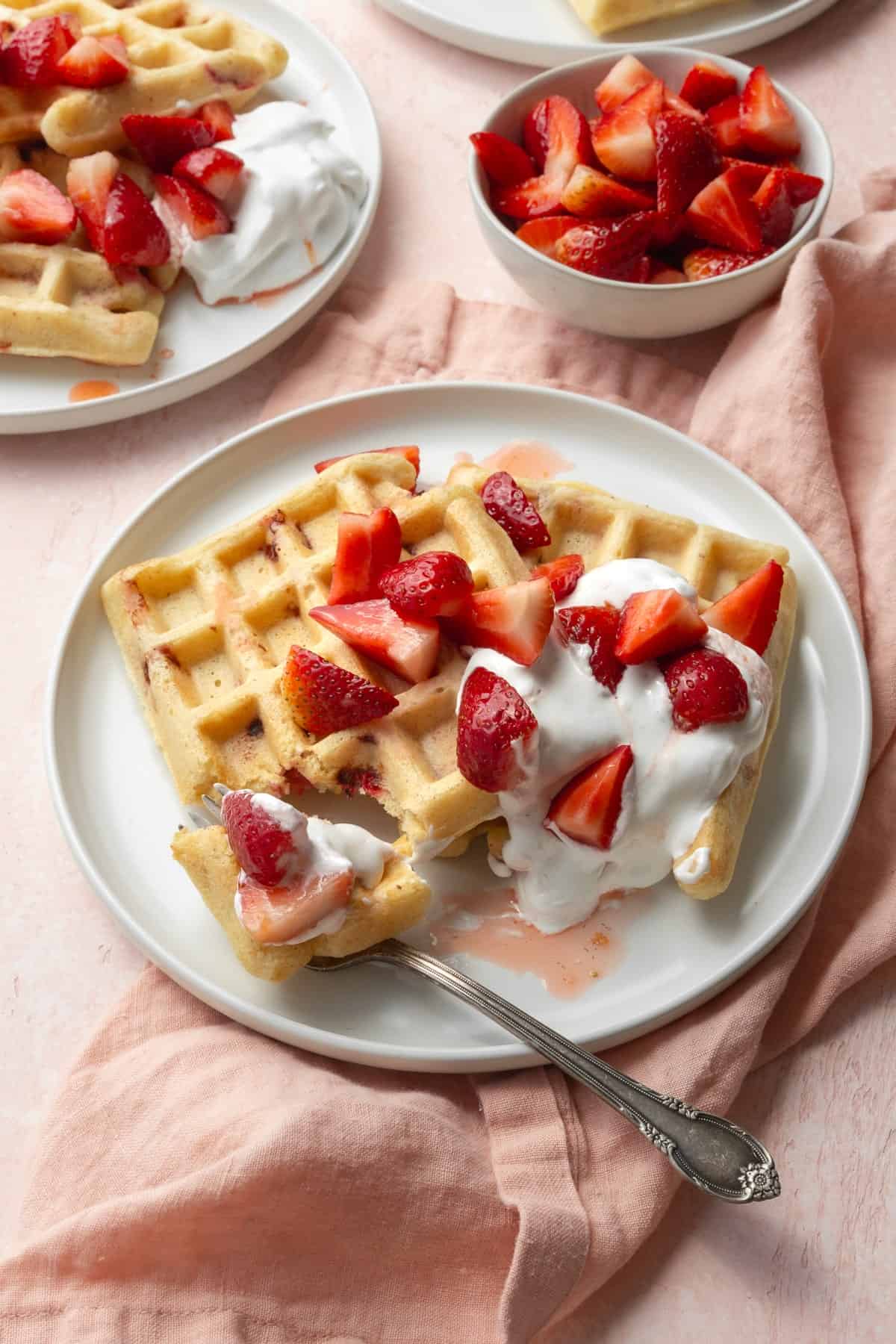 A white plate with strawberry waffles, with a silver fork resting on the plate with a bite of waffle on it.