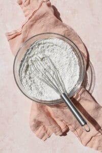 A glass bowl of dry ingredients whisked together on a pink surface.