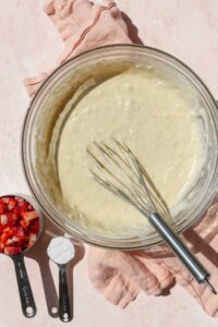 A bowl of waffle batter with strawberries and extra flour in measuring cups on the side.