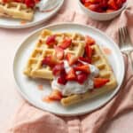 Two strawberry waffles on a white plate with whipped cream and sliced strawberries on top, on a pink napkin on a pink surface.