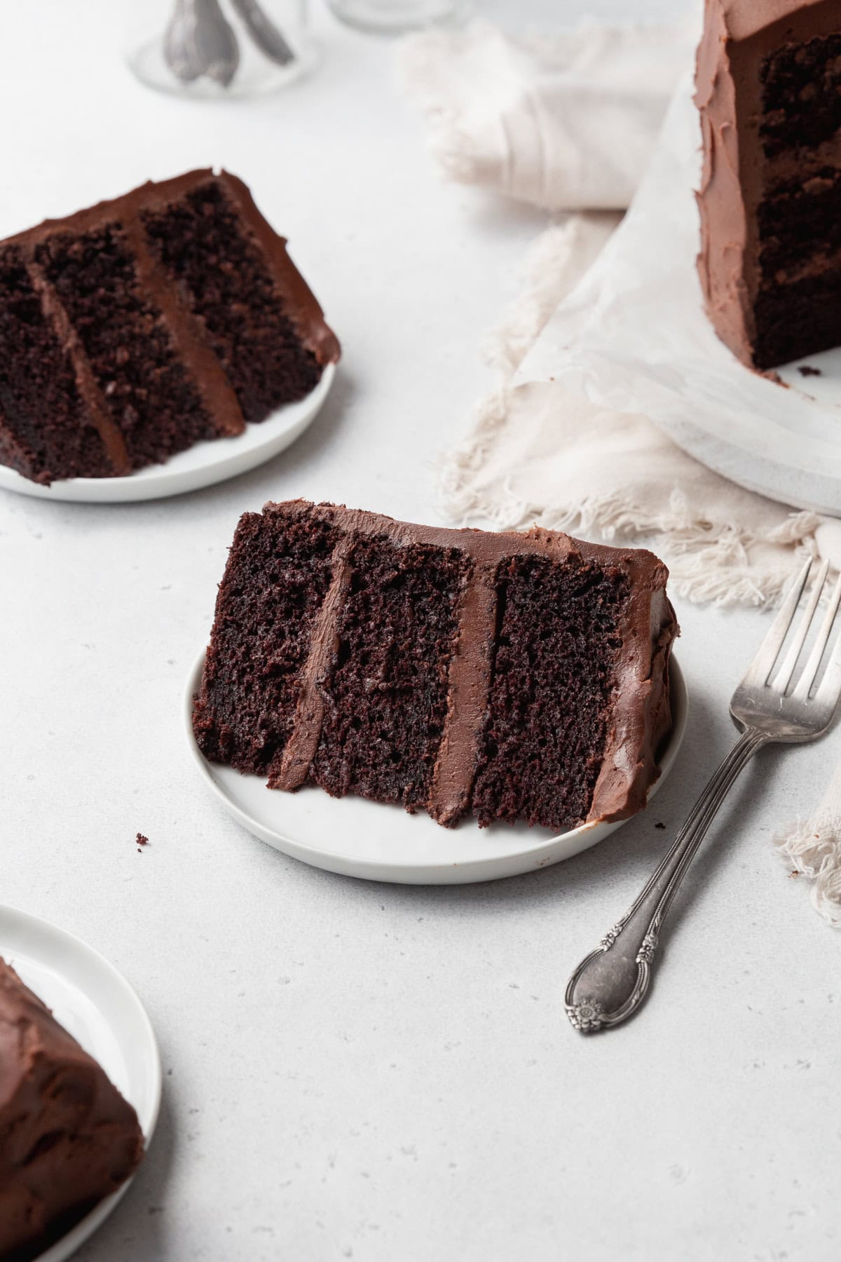 A slice of gluten-free chocolate cake on a small white plate with a silver fork next to it, and more cake slices around.