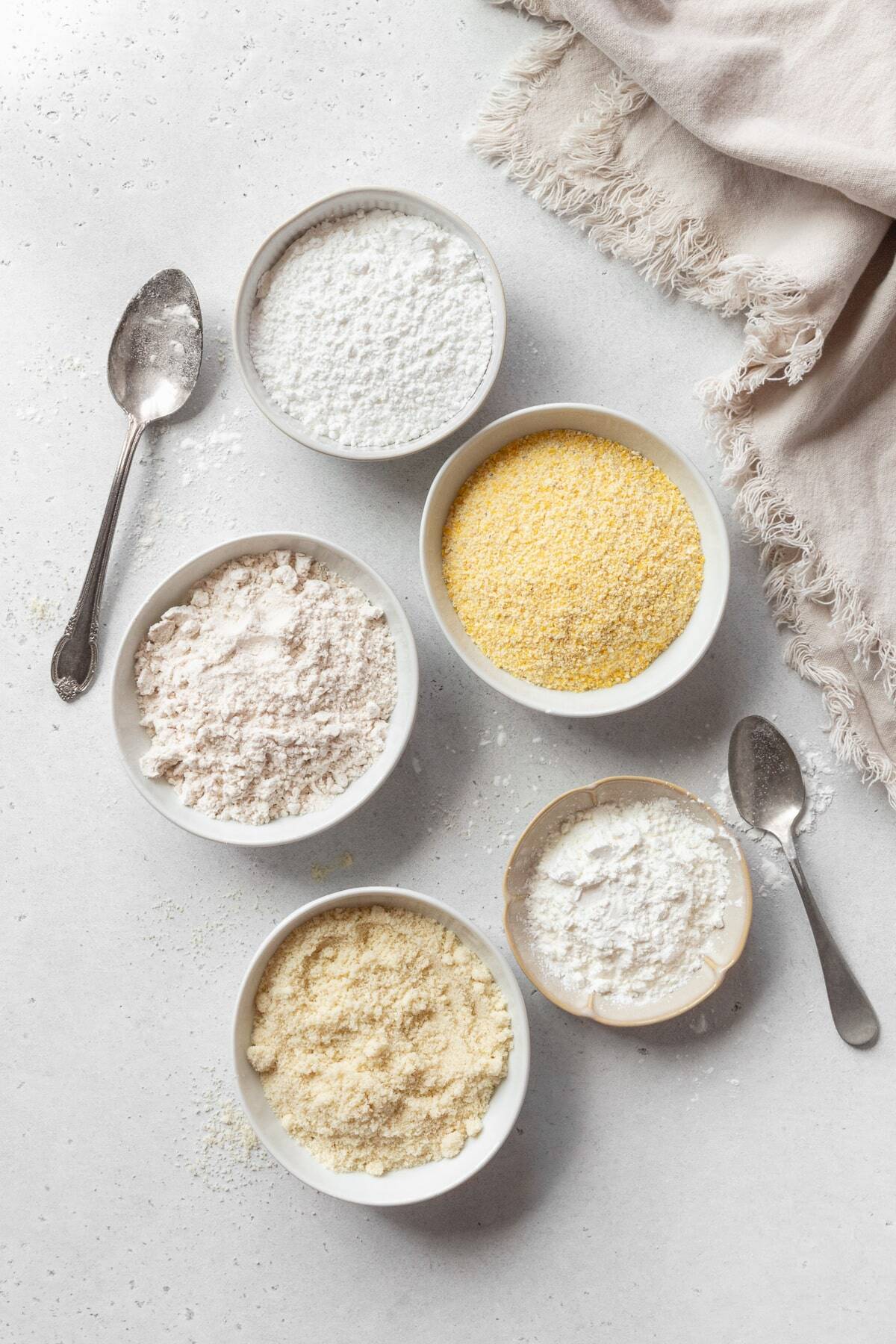 Bowls of gluten free flours on a white surface with a linen on the side.