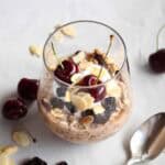 Overnight oats in a glass topped with yogurt, dried cherries, sliced almonds, and two fresh cherries on top.