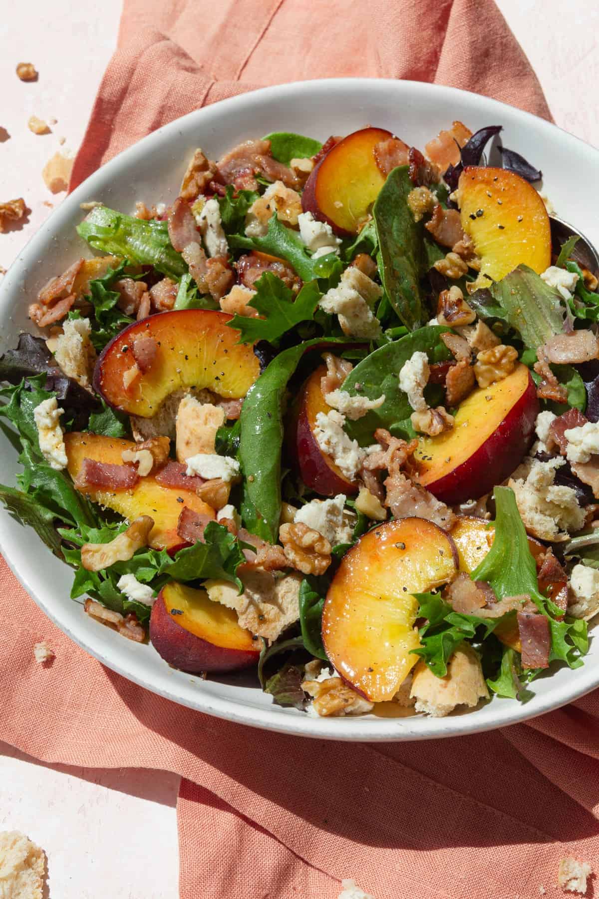 Peach salad with baby greens, cheese, and walnuts in a white bowl on a pink napkin.