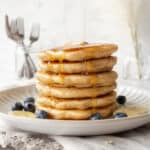 A stack of oatmeal pancakes on a plate with maple syrup being poured on top.