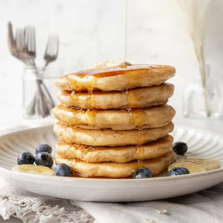 A stack of oatmeal pancakes on a plate with maple syrup being poured on top.
