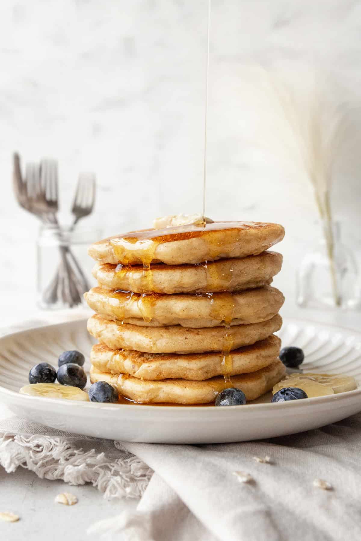 A stack of oatmeal pancakes and some blueberries on a plate with syrup being poured on top.
