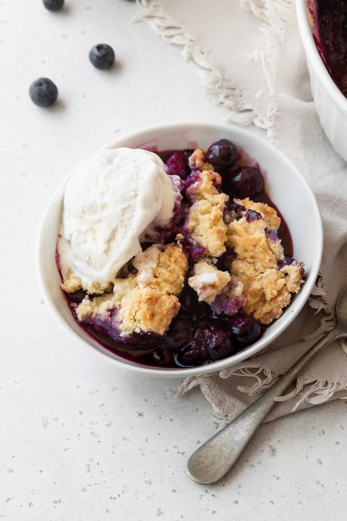 A bowl of blueberry cobbler with a silver spoon next to it on a white surface.